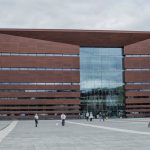 Nowoczesny gmach Narodowego Forum Muzyki we Wrocławiu z czerwonej bryły i przeszkloną fasadą, widziany z szerokiego placu, po którym spacerują mieszkańcy – symbol kosztownej, ale ważnej inwestycji kulturalnej miasta.