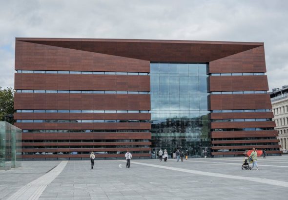 Nowoczesny gmach Narodowego Forum Muzyki we Wrocławiu z czerwonej bryły i przeszkloną fasadą, widziany z szerokiego placu, po którym spacerują mieszkańcy – symbol kosztownej, ale ważnej inwestycji kulturalnej miasta.