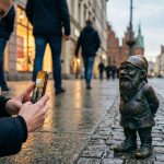 Turystka kuca na wrocławskim Rynku i fotografuje małego pomnikowego krasnala stojącego na mokrej kostce brukowej, symbol popularnej miejskiej gry w szukanie figurek.