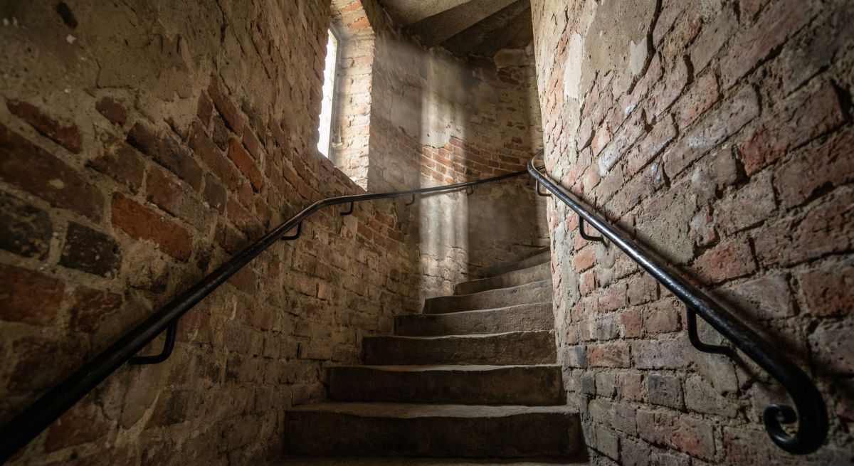 Kamienne, kręte schody w ceglanej wieży prowadzące na Mostek Pokutnic we Wrocławiu, oświetlone światłem wpadającym przez wąskie okno.