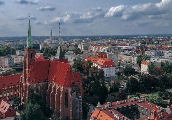 Panoramiczny widok na Wrocław z dominującą w kadrze kolegiatą św. Krzyża z czerwonym dachem i zieloną wieżą na Ostrowie Tumskim.