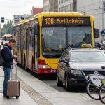 Przystanek w centrum miasta: żółty autobus linii 106 „Port Lotniczy”, obok taksówka i stojak rowerów miejskich, a pasażer z walizką sprawdza telefon.